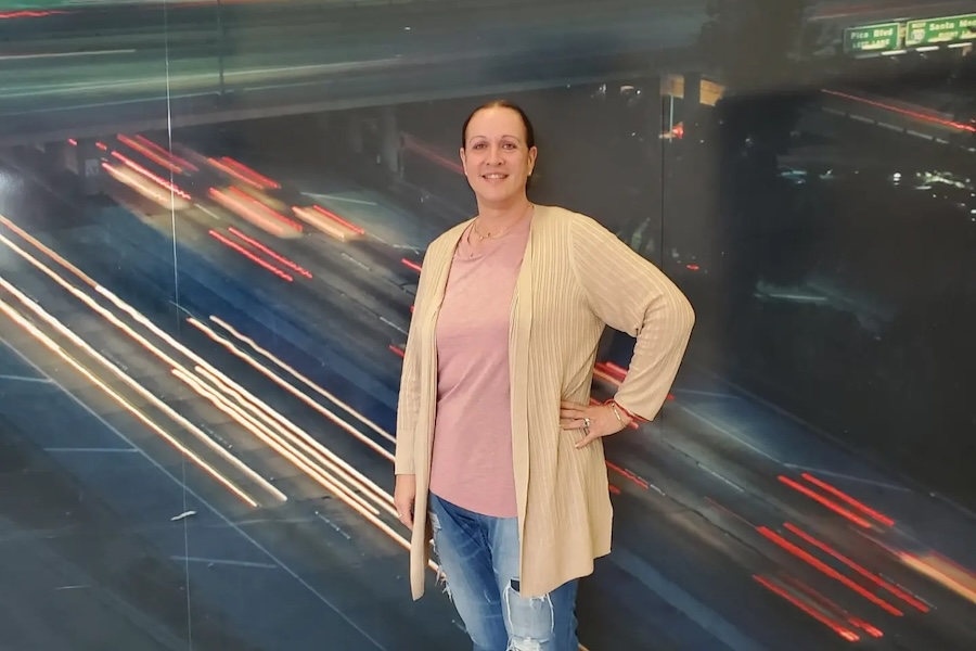 Yana Shmuliver, founder of K.A.U.S.E., in a long beige cardigan and jeans poses in front of a mural of highway light trails.
