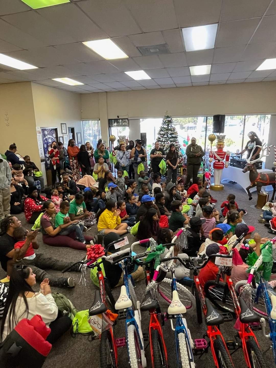 Large group of children and adults gathered at a holiday event indoors, with rows of colorful bicycles and a decorated Christmas tree in the background.