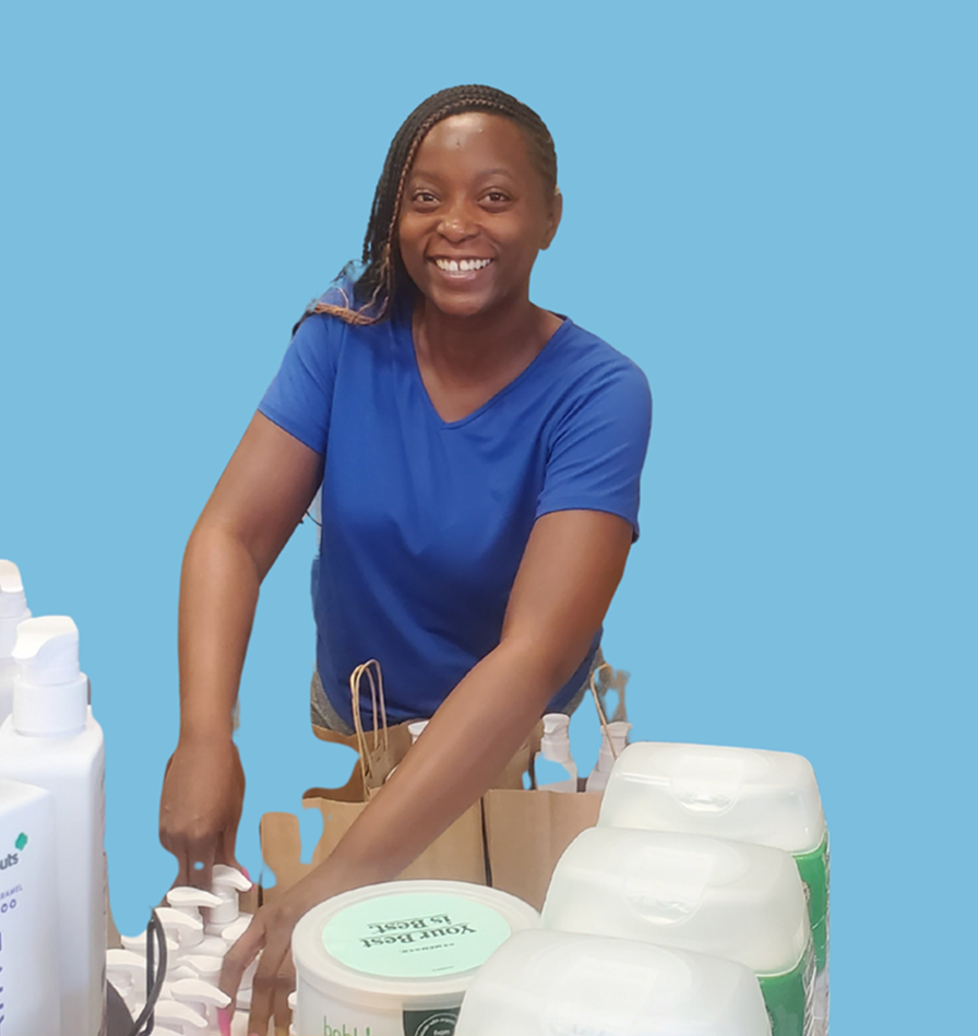 Smiling woman in a blue shirt reaching for hygiene products on a table, with a solid light blue background behind her.