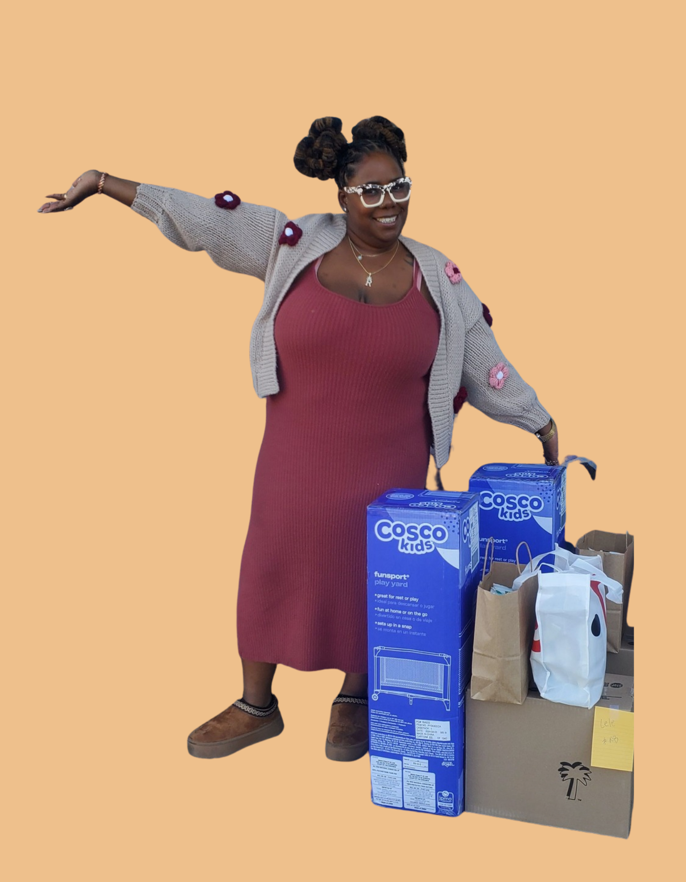Woman in a maroon dress and gray cardigan standing with arms outstretched in front of stacks of baby supplies including play yards and shopping bags.