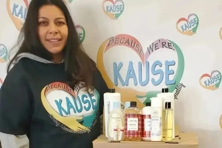 Woman wearing a KAUSE sweatshirt stands beside a table of personal care products in front of a KAUSE-branded backdrop.