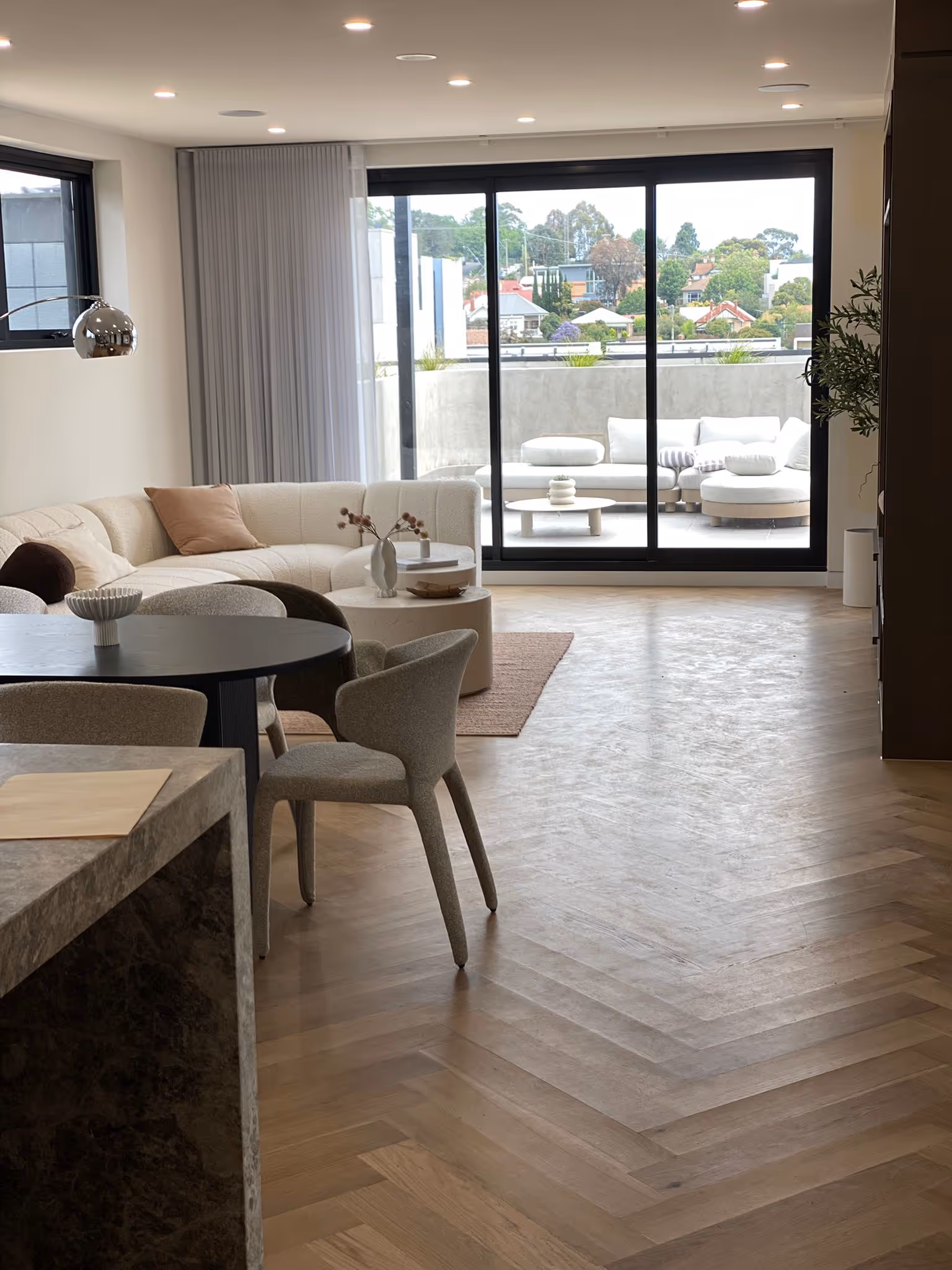 Modern living room with beige sectional sofa, round black dining table, gray chairs, and large sliding glass doors opening to outdoor seating area.