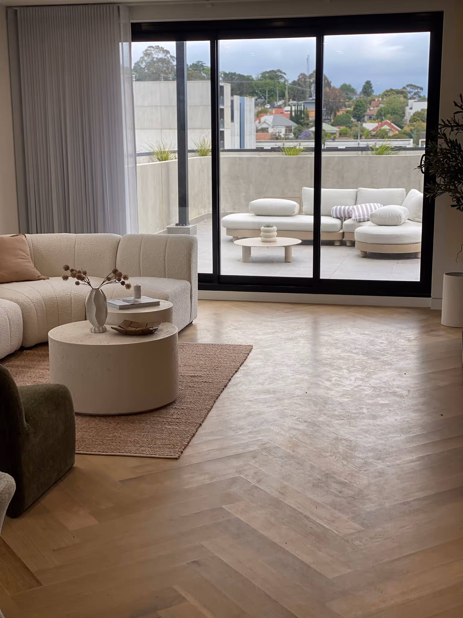 Modern living room with curved white sofa, round coffee tables on a rug, and sliding glass doors opening to a balcony with outdoor seating.