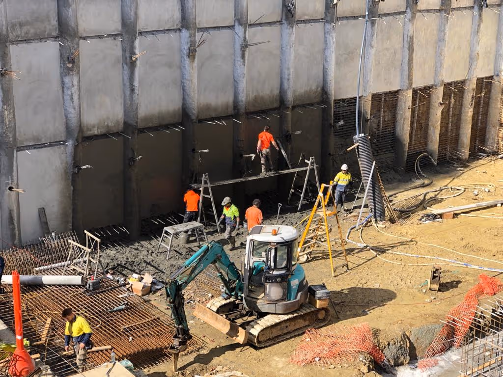 Construction site with workers in safety vests operating machinery and working near a large concrete retaining wall.
