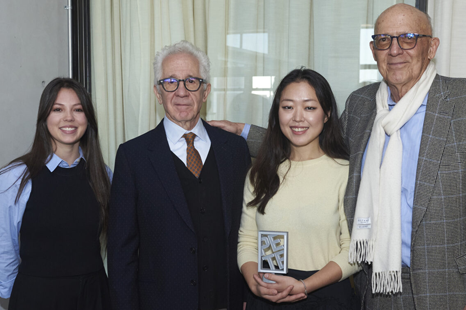 Two young women, one holding a small award plaque, pose with two older men in suits at the 2023 PETE Prize announcement.