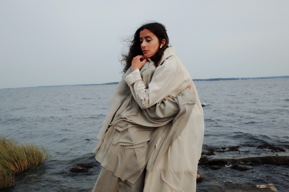 A model wrapped in a layered, pale gray oversized coat stands on a rocky shoreline with the sea behind her.