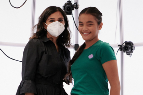 A young girl modeling a green Girl Scouts top stands beside a woman wearing a face mask on a photo shoot set.