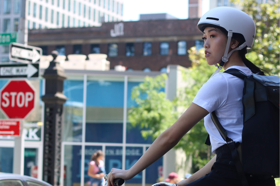 A young woman in a white helmet and backpack rides a bike through a city street, glancing over her shoulder.
