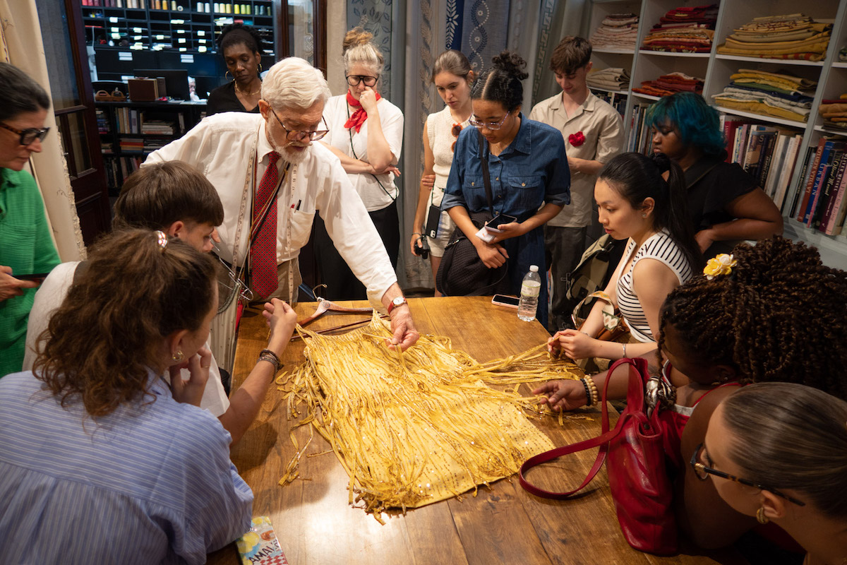 About a dozen young people crowd around a large wooden table in a workroom whose walls are lined with shelves of folded fabrics, stacked trims and books. A white-haired man in a white shirt, red patterned tie and glasses leans over the table and holds up a wide fringed panel of golden-yellow threads, turning it toward the group as they lean in to look.