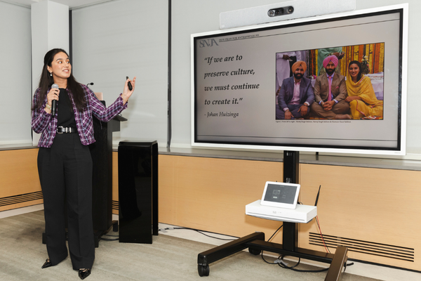A young woman in a purple plaid jacket, black top and wide black trousers stands in a pale classroom, holding a microphone in one hand and raising the other as she speaks and looks toward a screen showing a presentation slide with a photograph of three men in turbans seated side by side.