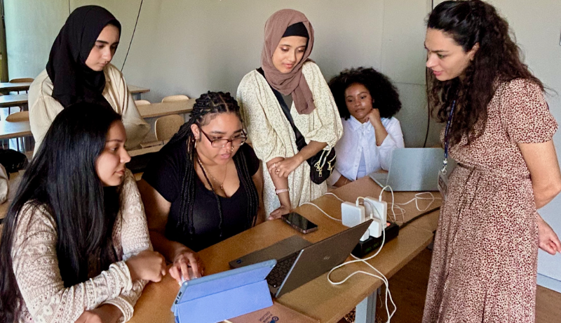 Six women gather around a pale wooden table in a bright classroom, all looking down at an open silver laptop and a tablet propped on a stand. A woman in a brown and white floral print dress stands at the right, leaning in toward the screen as she explains something.