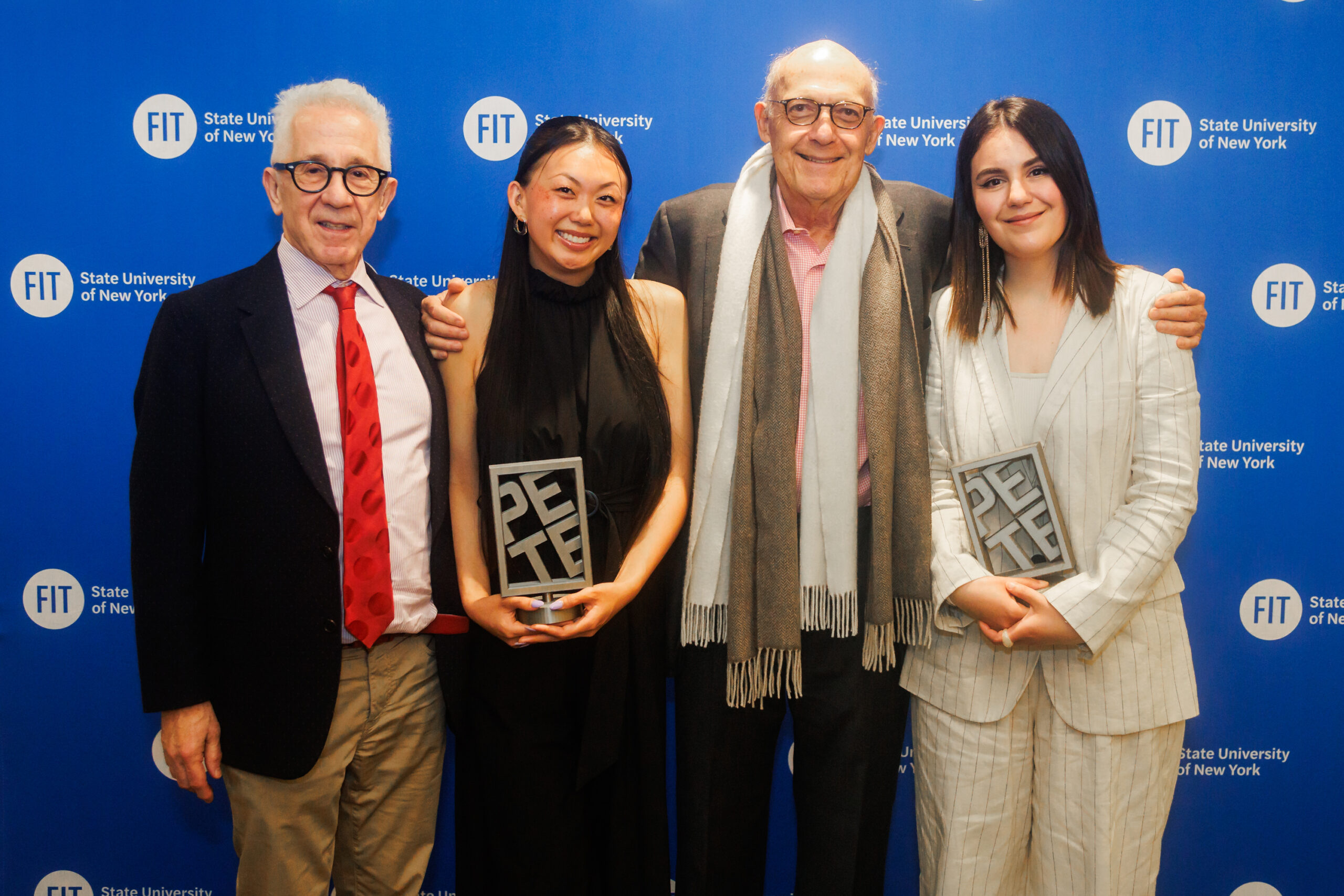 Four people stand shoulder to shoulder in front of a royal blue step-and-repeat backdrop tiled with the round FIT logo. Two young women each hold a chunky silver trophy whose blocks spell PETE, flanked by two older men in jackets, everyone smiling at the camera.