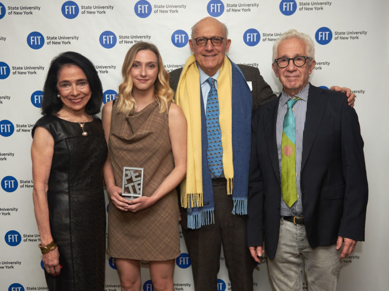 Four people stand shoulder to shoulder in front of a step-and-repeat backdrop printed with blue FIT logos. From left: a woman in a black sleeveless dress; a young blonde woman in a tan plaid shift dress holding a small clear glass award block; an older man in a tweed jacket with a long fringed scarf; and an older man in a navy blazer and striped shirt.