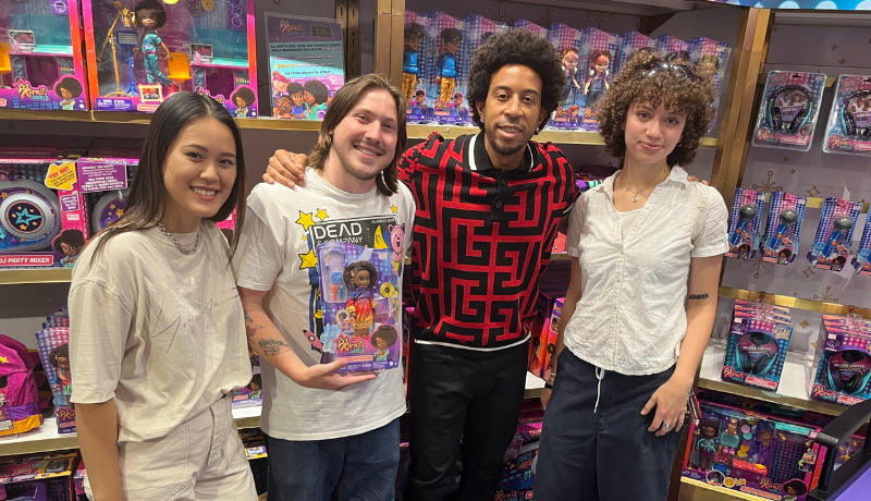 Four people pose together inside a toy store, in front of gold-trimmed shelves packed with brightly boxed dolls and accessories in pink and purple packaging. One man holds up a boxed doll set; another wears a red and black geometric-print jacket.