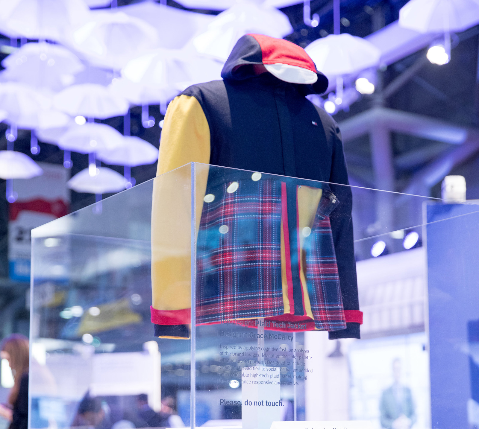 A hooded jacket displayed on an invisible form inside a clear acrylic case at a trade show. The body is navy, the left sleeve is pale yellow, the hood lining and hem trim are red, and a large red, blue and white plaid panel covers the front. Rows of upside-down white umbrellas hang from the ceiling under blue lighting.