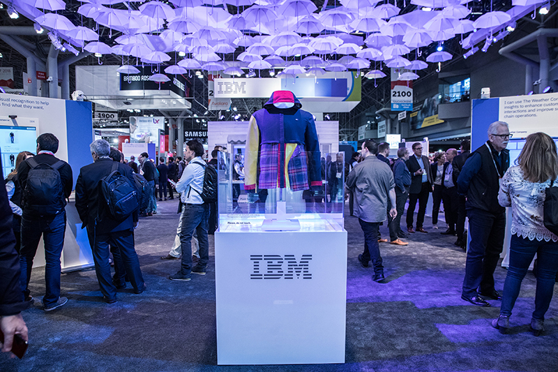 A busy trade show floor under a ceiling densely hung with hundreds of pale upside-down umbrellas lit purple. In the center, a hooded jacket with a pink hood lining, yellow sleeve and a red and blue plaid front panel stands inside a clear display case on a white plinth marked with the striped IBM logo.