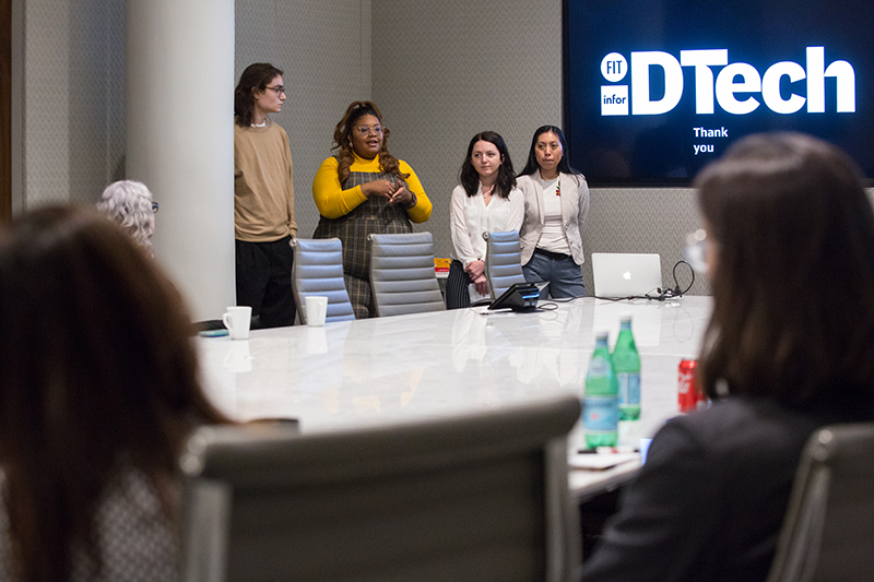 Four young women stand together at the head of a long glossy white conference table in a dimly lit corporate meeting room. On the wall behind them a large screen displays a white-on-navy logo reading FIT infor DTech, with the words Thank you underneath.