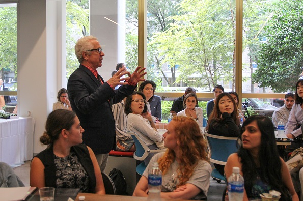 An older man with swept-back white hair, round tortoiseshell glasses and a patterned red tie stands at the left, gesturing with both hands raised as he speaks to a seated audience of roughly twenty young adults in a bright room with floor-to-ceiling windows looking onto green trees.