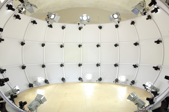Interior view of a large white domed photography studio. Curved white panels form the walls, and dozens of small black cameras are mounted in evenly spaced rows across them, interspersed with silver floodlights angled inward. The floor at the center is bare pale wood.
