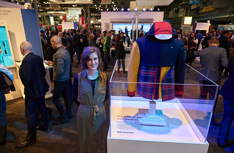 A crowded trade show floor. In the foreground a young woman in a grey belted shirtdress stands smiling beside a clear acrylic display case holding a jacket with a red hood, royal blue body, one yellow sleeve, and a red and blue plaid front panel. Dozens of attendees fill the aisles behind her.