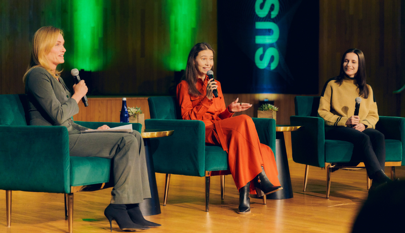 Three women sit in green velvet armchairs on a wooden stage, two of them holding microphones mid-conversation, with small plants on gold side tables and green stage lighting against a dark screen behind them.