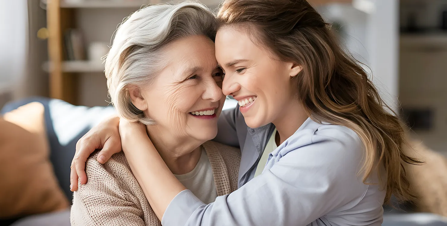 Young woman embraces and smiles warmly with an elderly woman in a cozy living room.