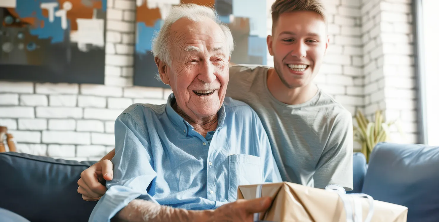 Smiling elderly man holding a wrapped gift with a young man hugging him from behind on a couch.