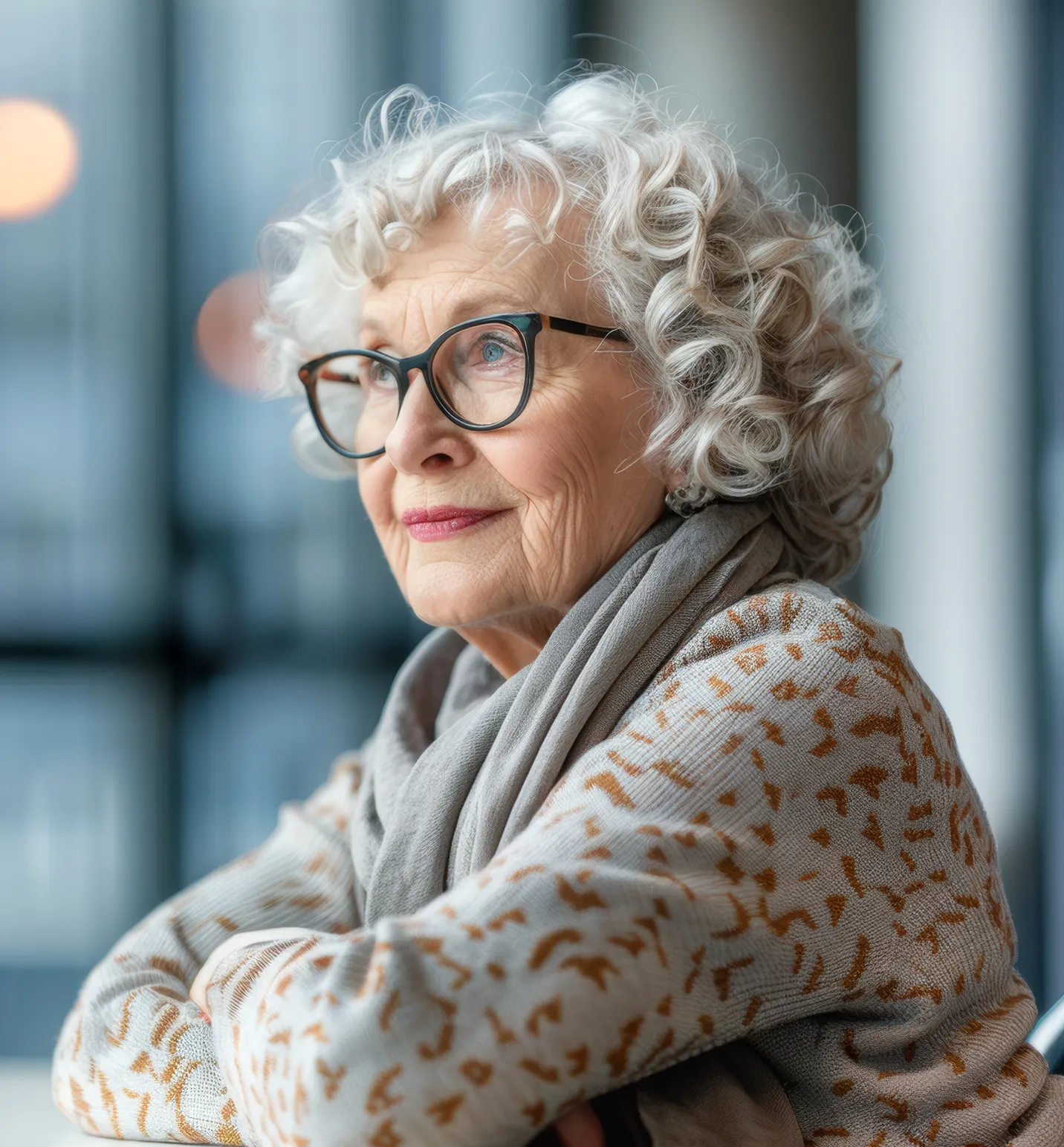 Elderly woman with curly white hair and glasses wearing a patterned sweater and gray scarf looking thoughtfully into the distance.