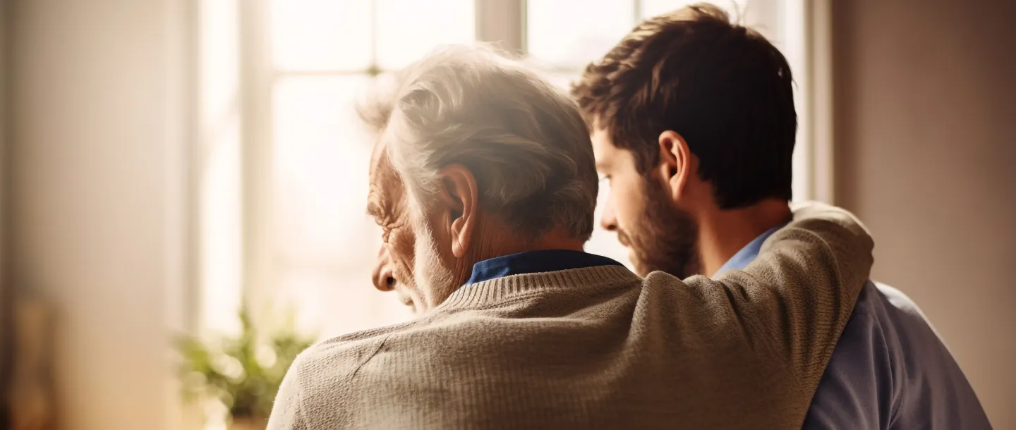 Older man with gray hair and a younger man with beard embracing while looking out a window indoors.