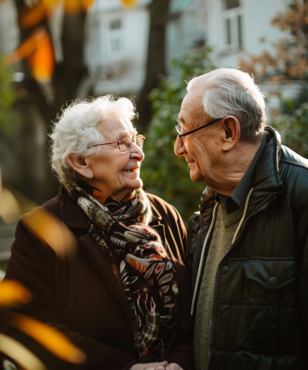 Elderly couple wearing glasses warmly smiling at each other outdoors amidst autumn foliage.