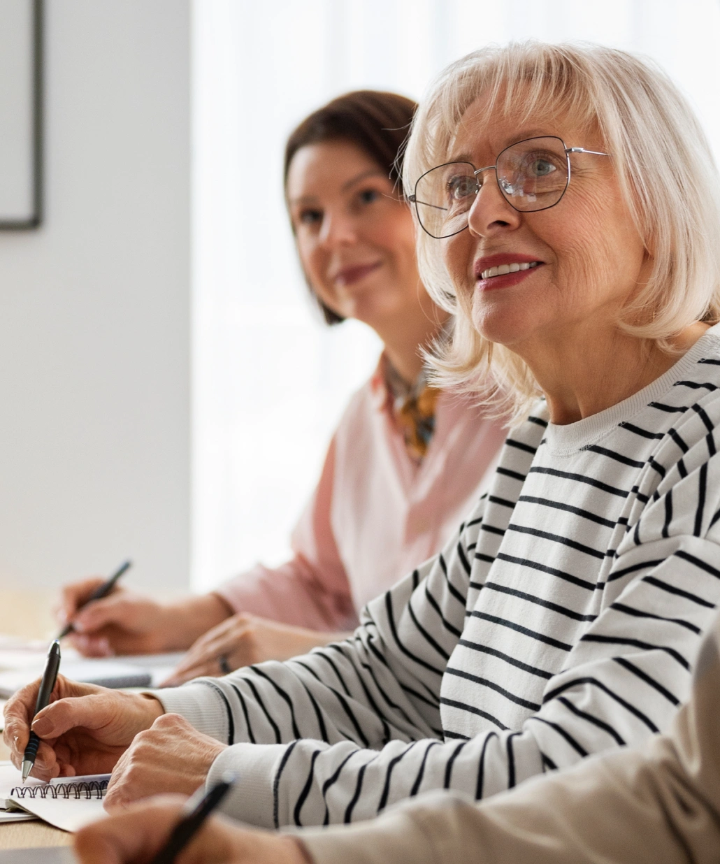 Older woman with glasses and striped shirt smiling while writing in a notebook at a table with others.