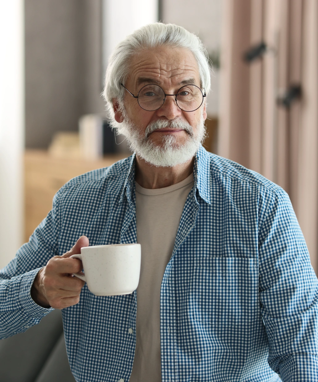 Elderly man with white hair and beard wearing glasses and a blue checkered shirt holding a white mug.