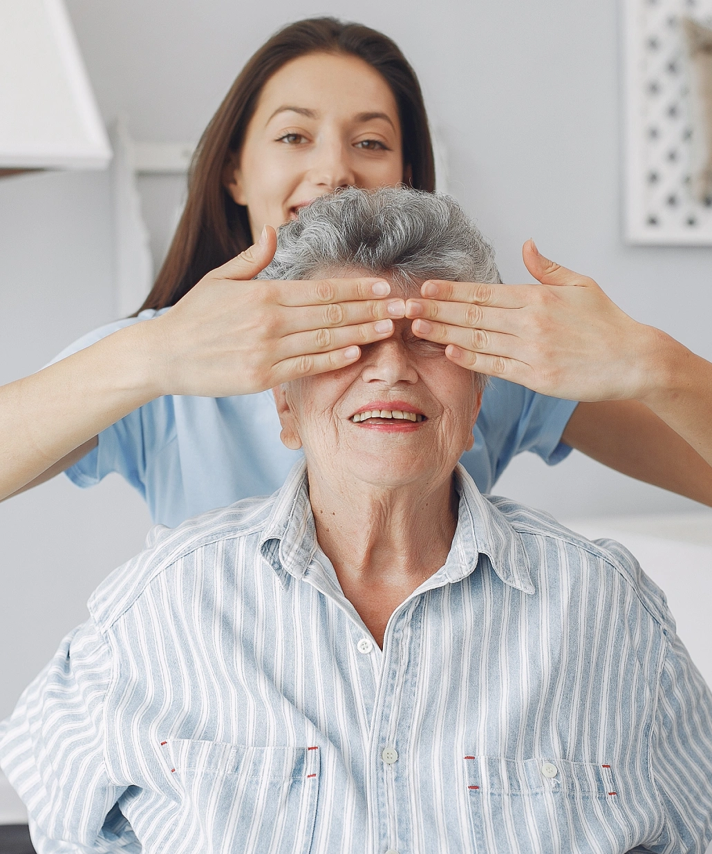 Young woman playfully covering the eyes of a smiling elderly woman wearing a striped shirt.