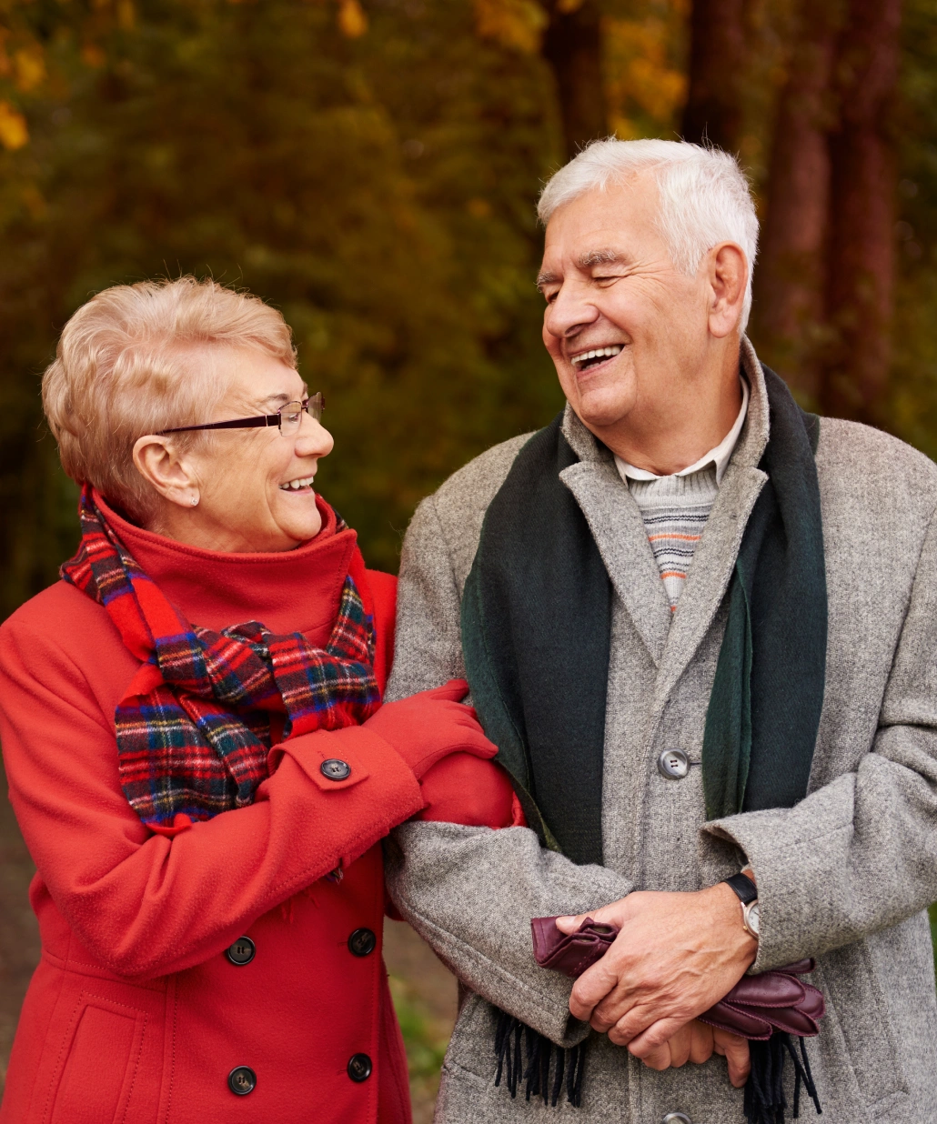 Smiling elderly couple dressed in autumn coats and scarves, walking arm in arm outdoors with trees in the background.