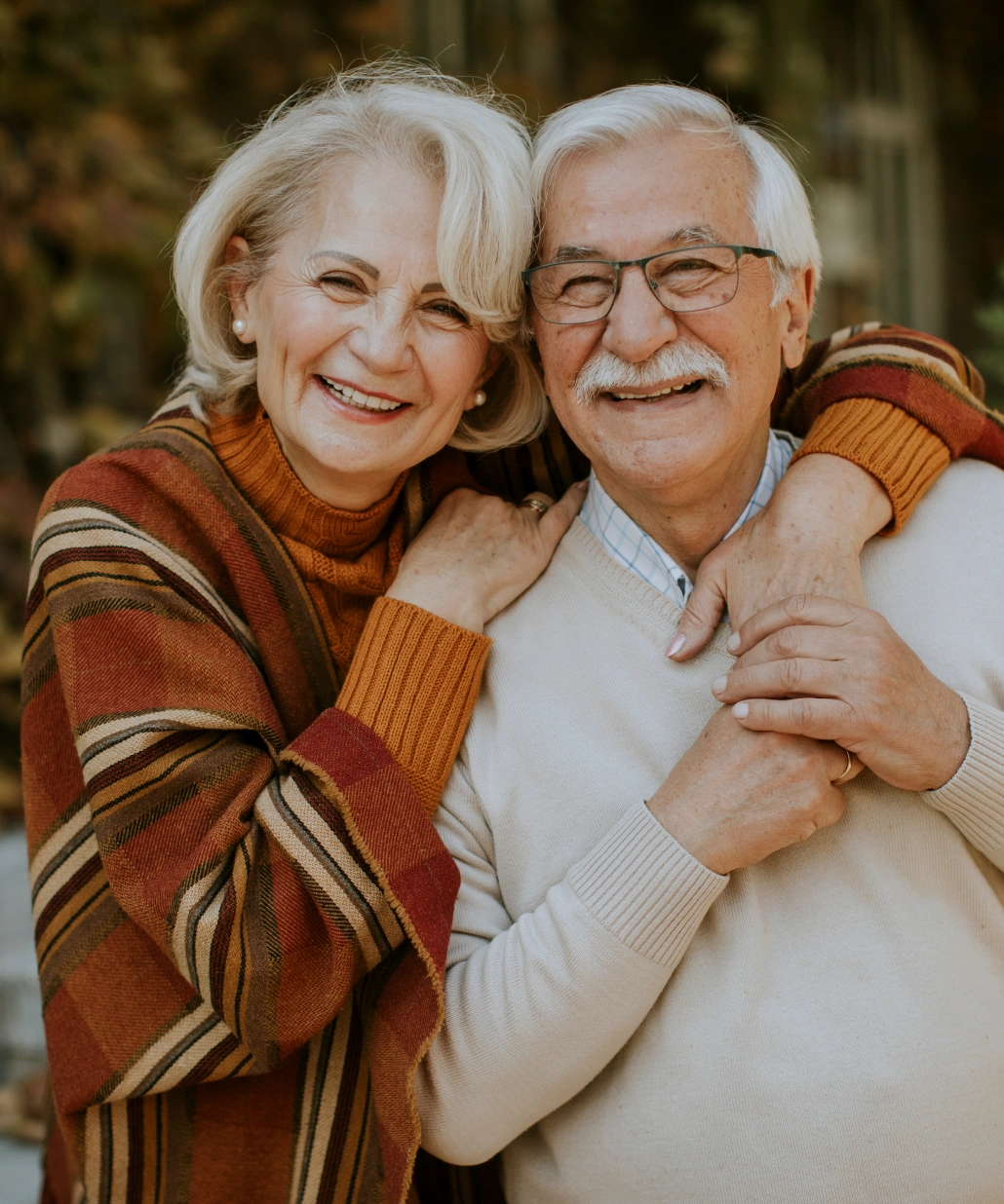 Smiling elderly couple embracing outdoors, the woman wearing a striped shawl and the man in a cream sweater.