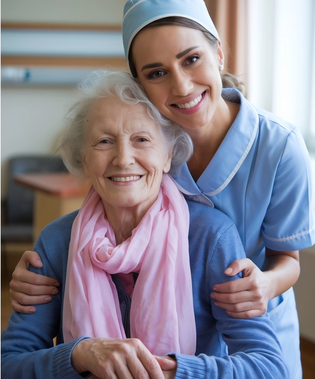 Smiling elderly woman wearing a pink scarf embraced by a smiling nurse in blue uniform.