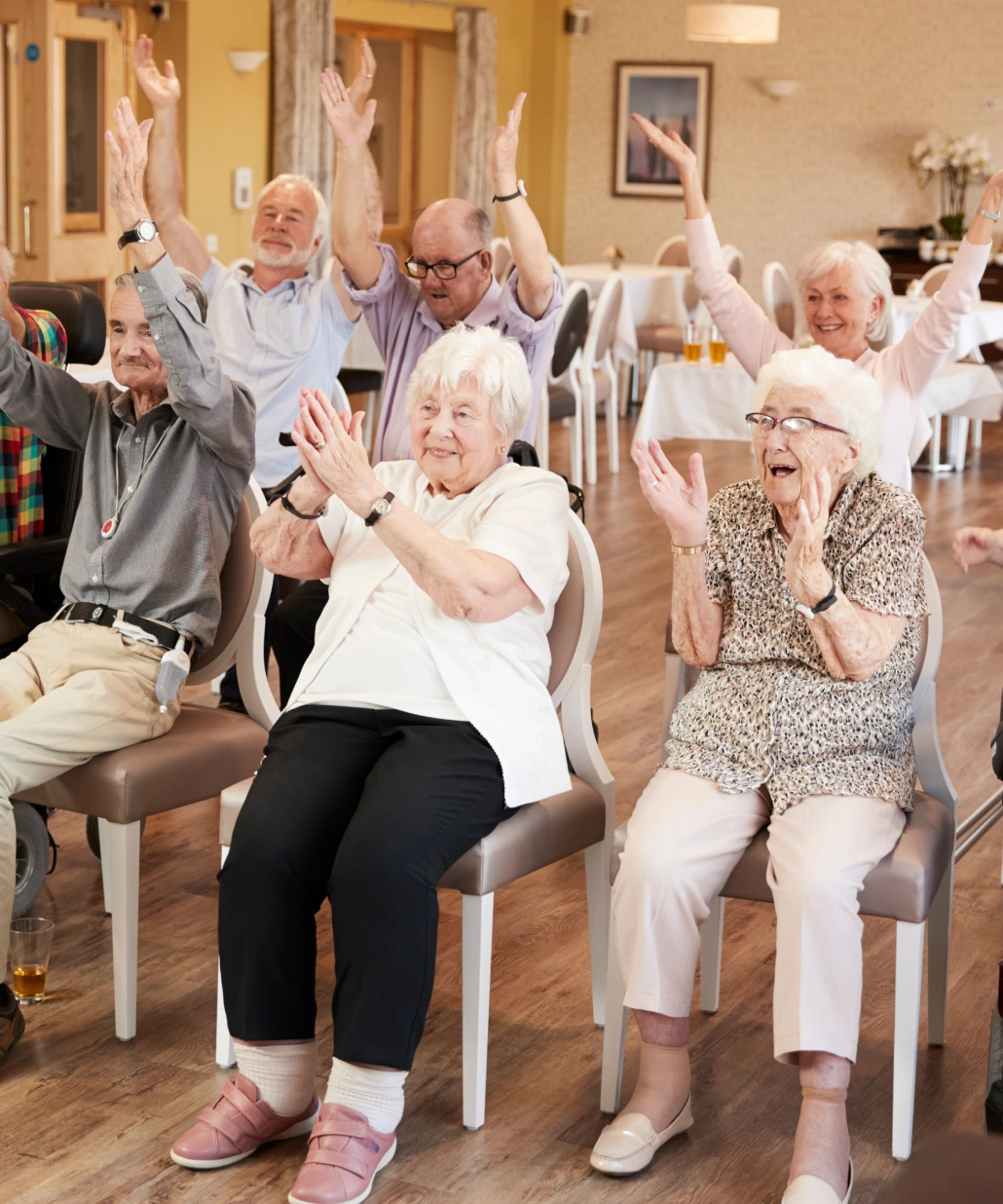 A group of elderly people seated on chairs in a room, happily raising their arms and clapping during a group activity.