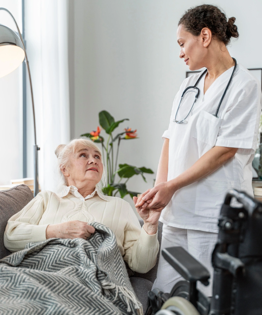 Nurse in white uniform holding hands and comforting an elderly woman sitting on a couch with a blanket.