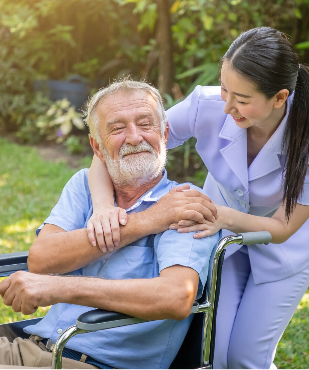 Smiling elderly man sitting in a wheelchair outdoors, embraced and supported by a female caregiver in a white uniform.