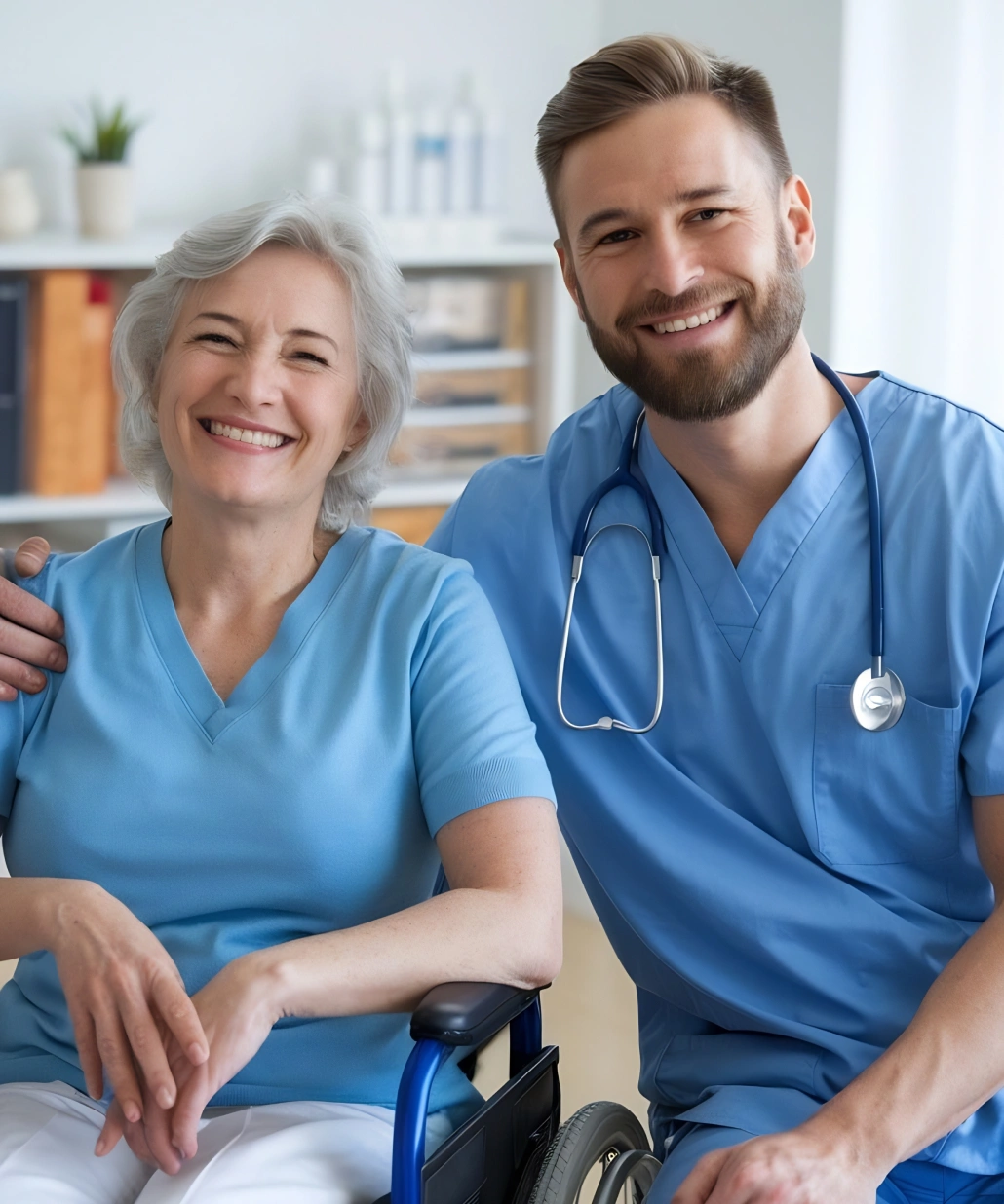 Smiling elderly woman in a wheelchair with a male healthcare professional wearing blue scrubs and a stethoscope.