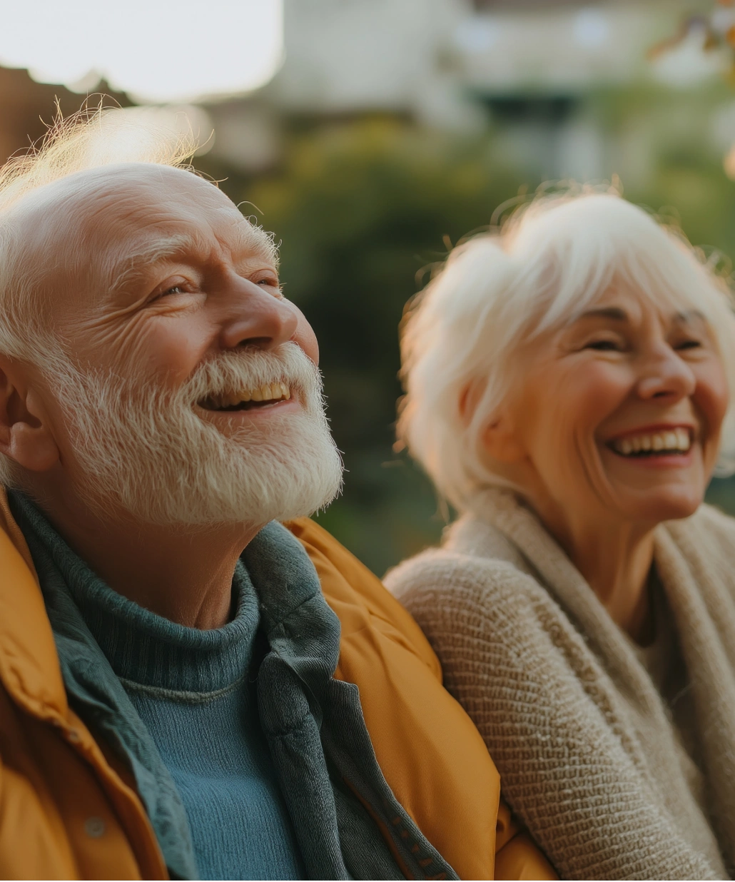 Elderly man with white hair and beard wearing a yellow jacket smiling outdoors next to a smiling elderly woman in a beige sweater.
