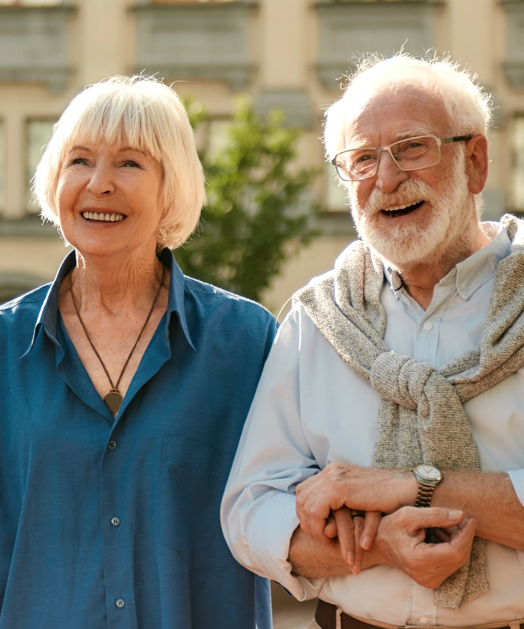 Elderly couple smiling outdoors, the man wearing glasses and a sweater over his shoulders, the woman in a blue shirt with a necklace.