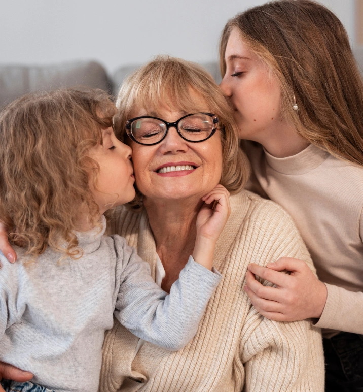 Smiling elderly woman wearing glasses receiving kisses on both cheeks from two girls in light-colored sweaters.