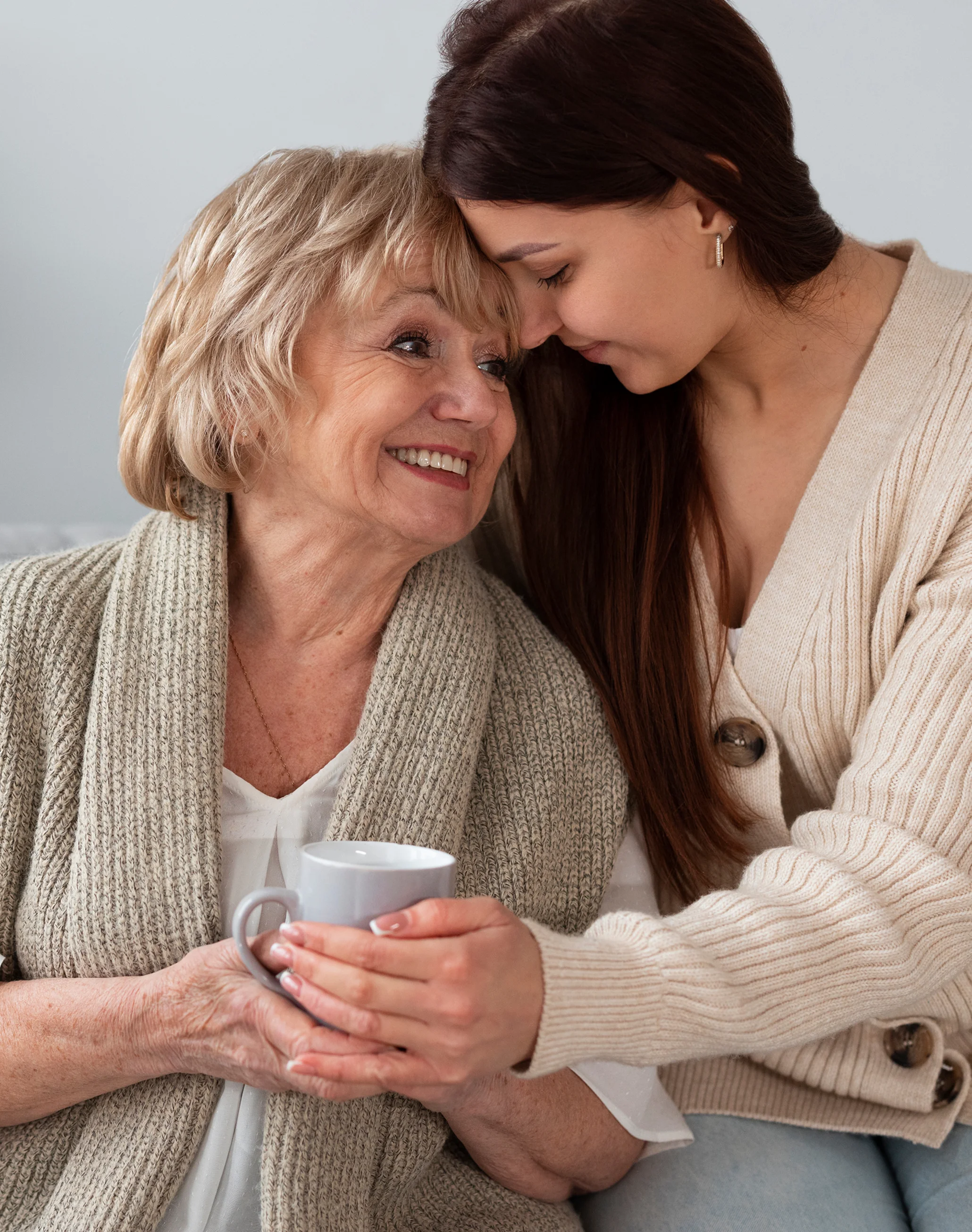 Smiling elderly woman holding a mug with a younger woman hugging her and resting their foreheads together.