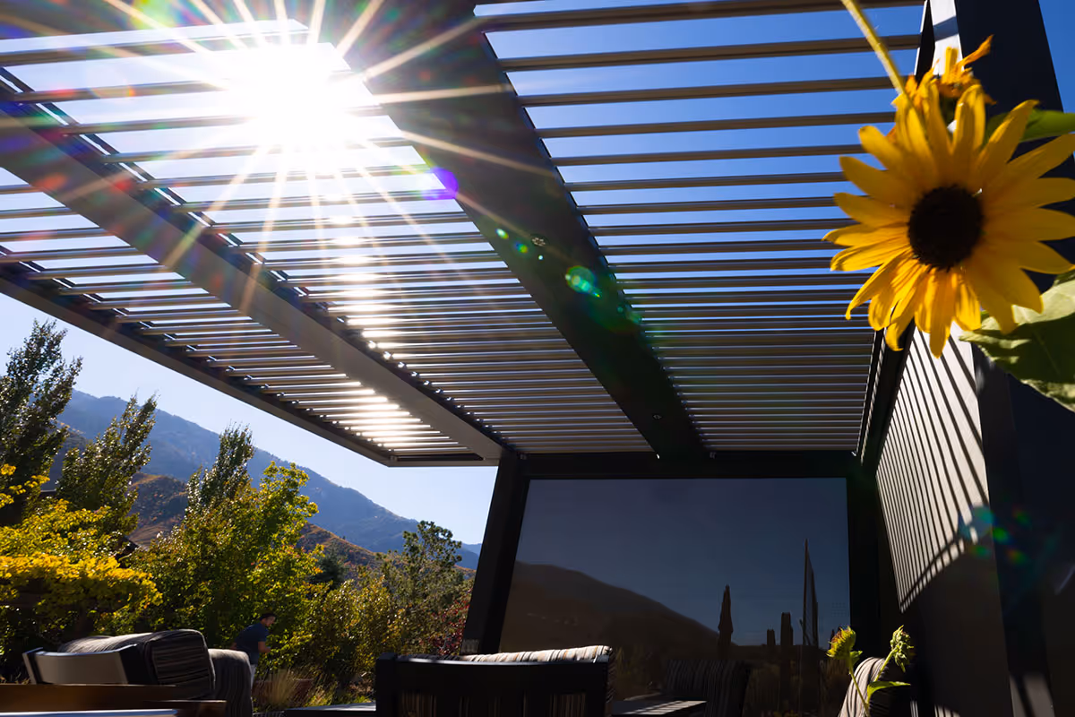 Sunlight peeking through the roof of a motorized pergola with one side that is utilizing a motorized shade.