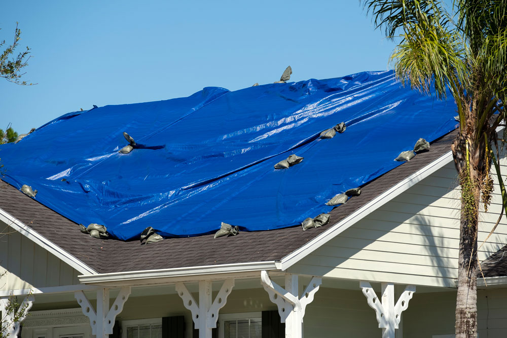 A roof covered with a blue tarp that has been damaged after a hurricane