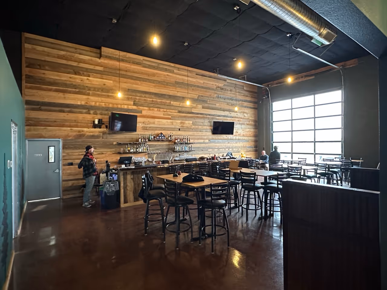 Interior of a modern bar with wooden walls, hanging lights, two TVs, and several tables and high chairs, with a large window letting in natural light.