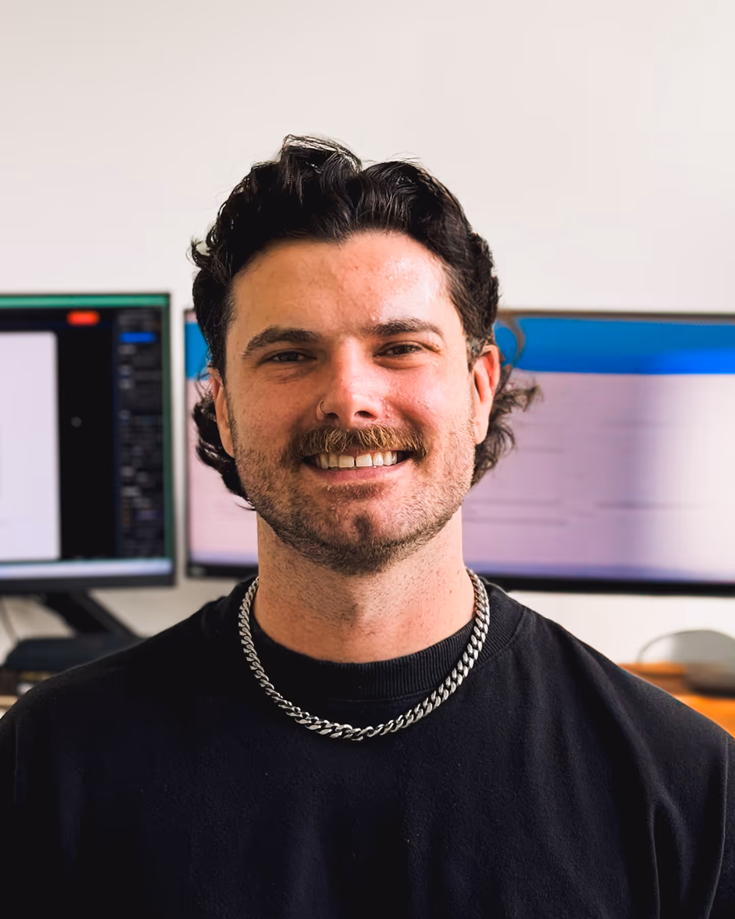 dark haired man with a mustache sitting in front of a desk with two monitors on it
