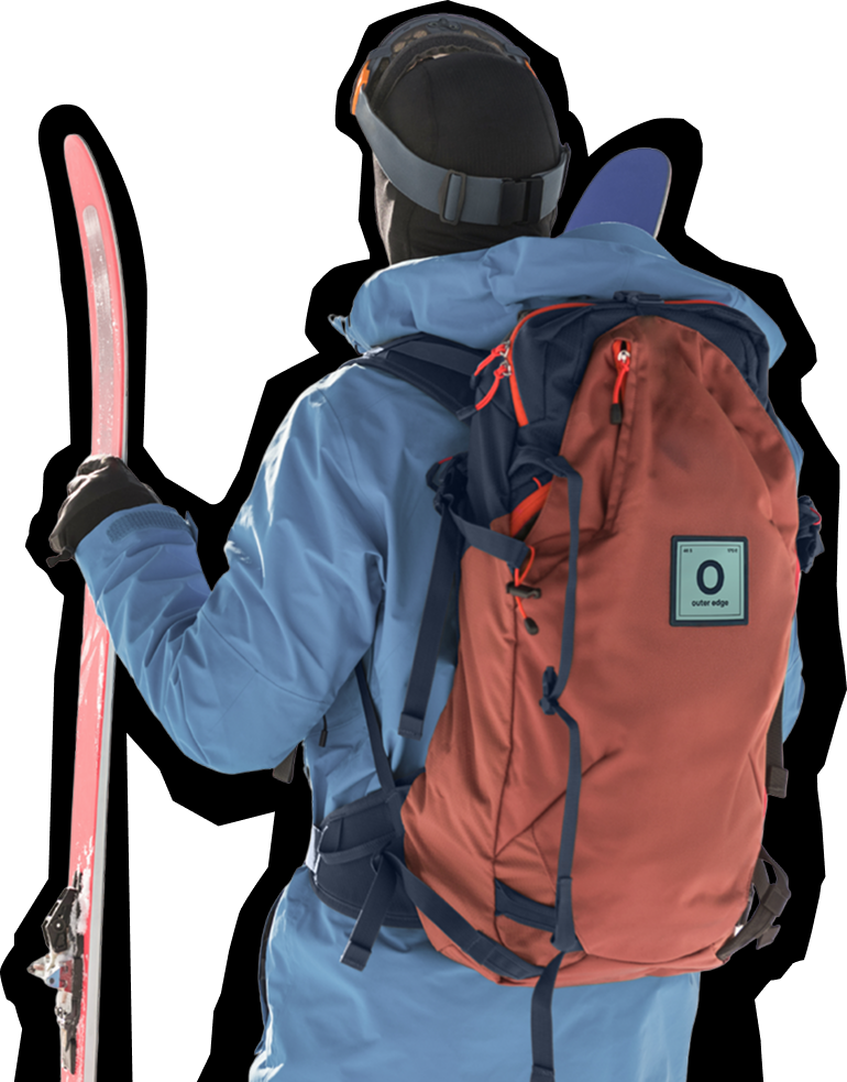 A person wearing a blue winter jacket and dark helmet stands with their back to the camera, holding a pair of skis. They are carrying a large orange and navy blue backpack with a small label or patch visible on it. The person appears to be equipped for backcountry skiing or ski touring, photographed against a light gray background.