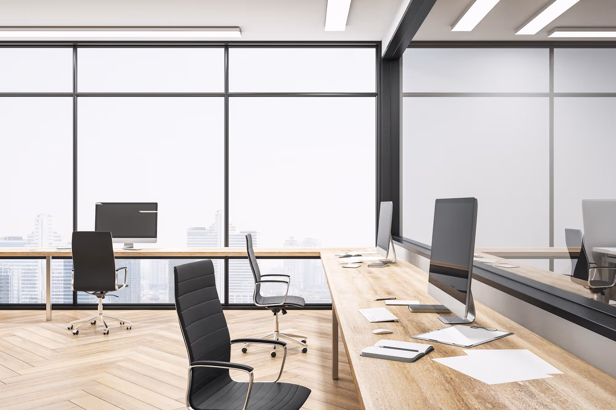 Modern office space with wooden desks, black ergonomic chairs, desktop computers, and large windows overlooking a cityscape.